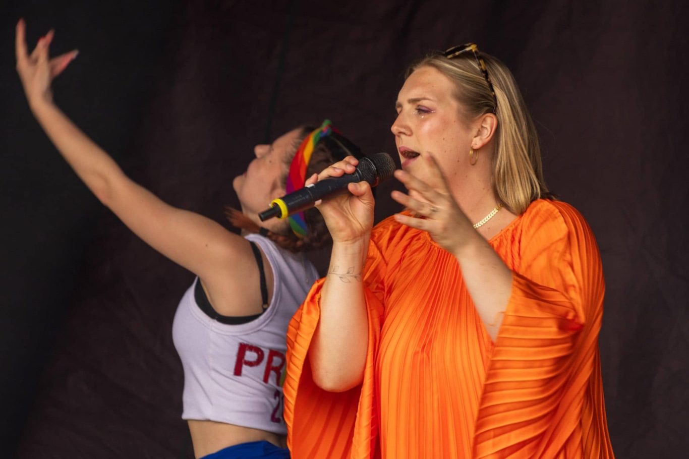 GYG - 2024 A woman in orange sings into a microphone while another dancer poses behind her.