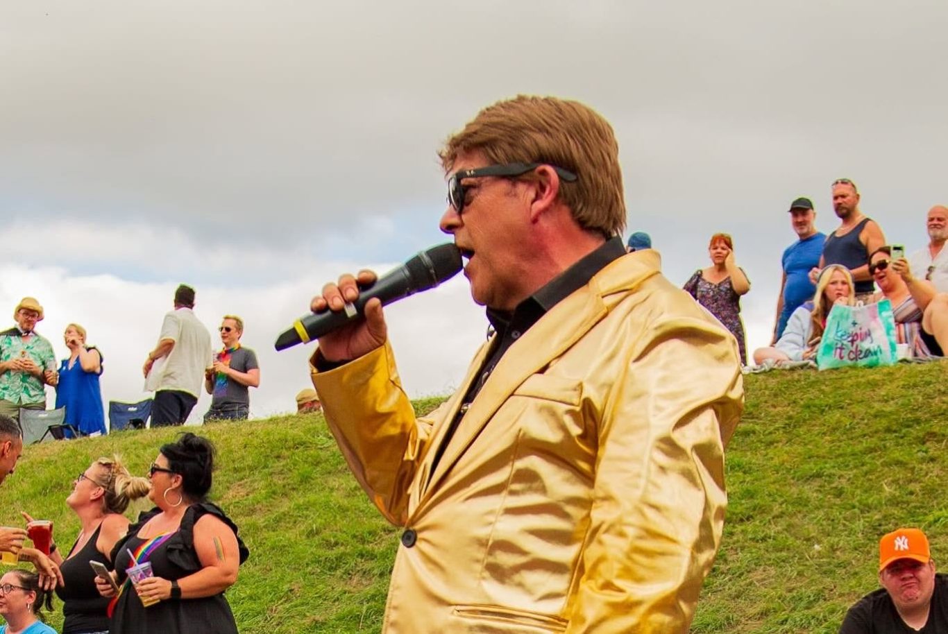 Elton John Tribute A man in a gold suit singing into a microphone at an outdoor event.
