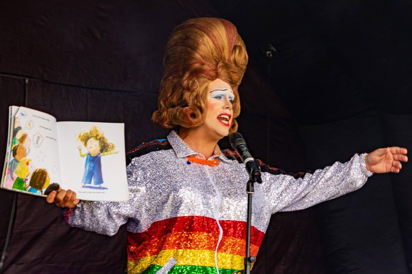 Mama G - 2024 A drag performer in a rainbow outfit reading a children's book on stage.