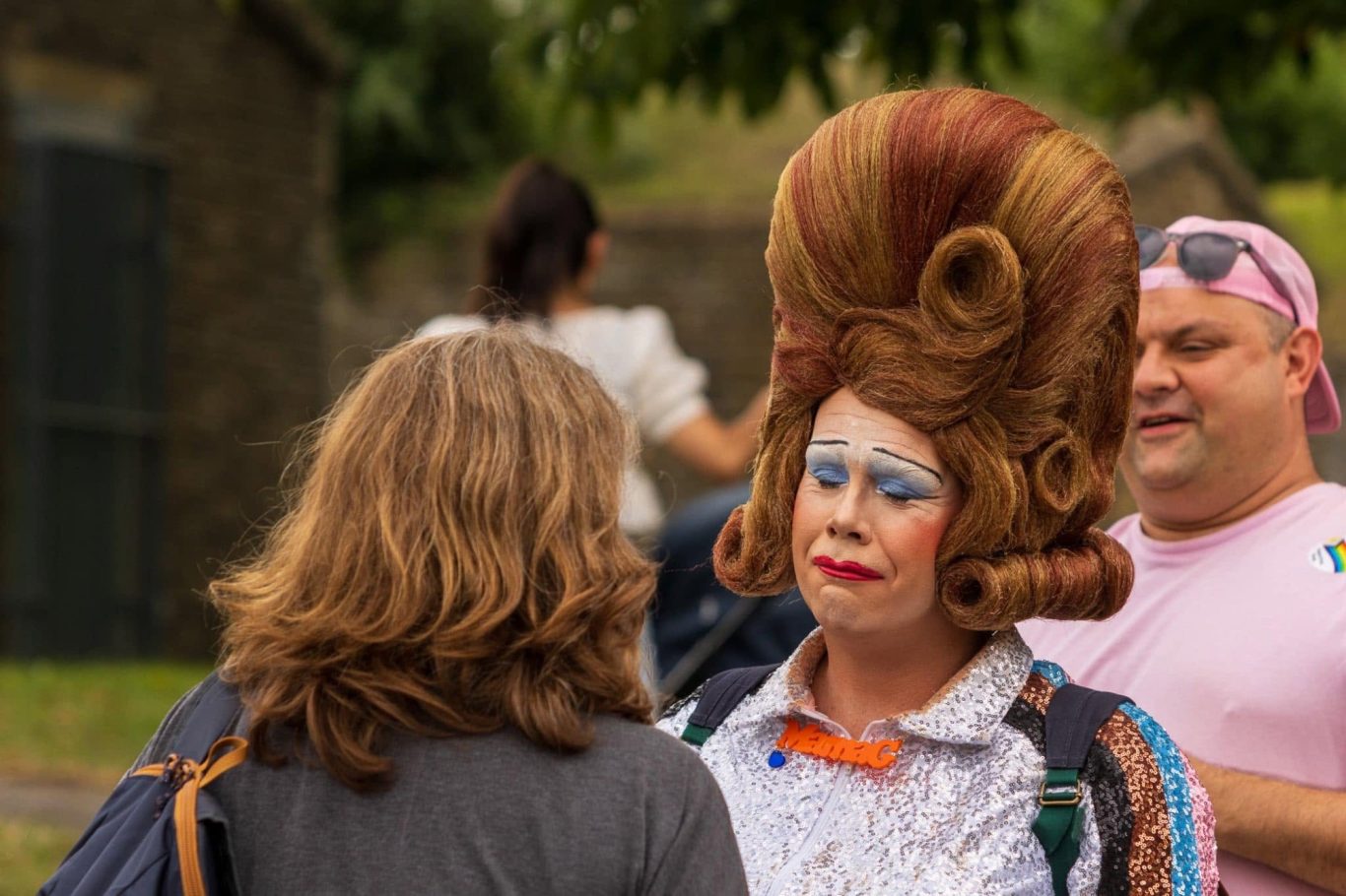 A drag performer in a flamboyant outfit converses with a woman outdoors.