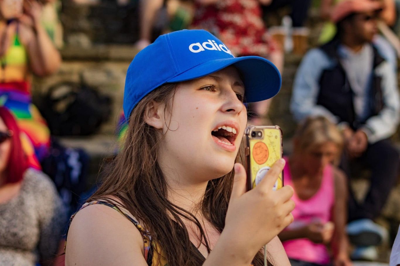 Young woman wearing a blue cap, singing and holding a phone, with a colourful crowd behind.