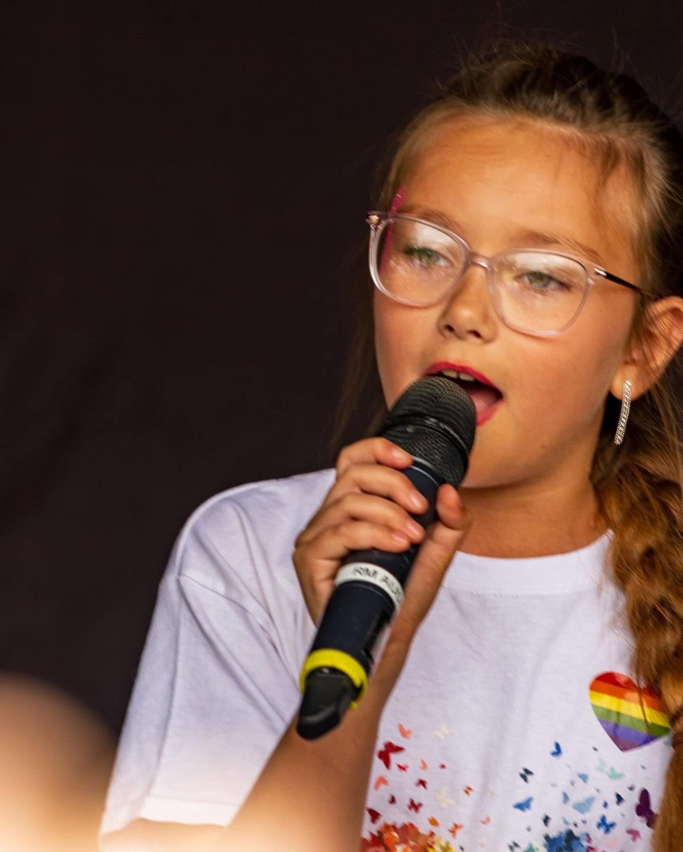 GYG - 2024 Young girl with glasses singing into a microphone, wearing a white shirt with a rainbow heart.