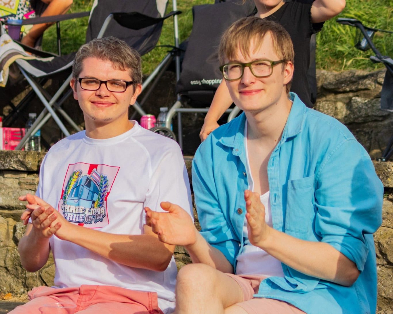 Two young adults sitting outdoors, smiling and clapping, wearing casual summer clothing.