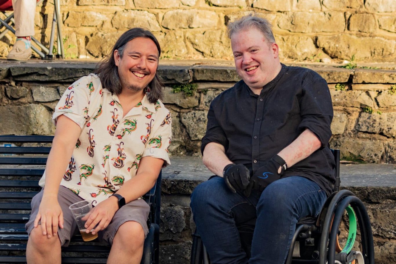 Two men smiling while seated on a bench, enjoying a casual moment outdoors.