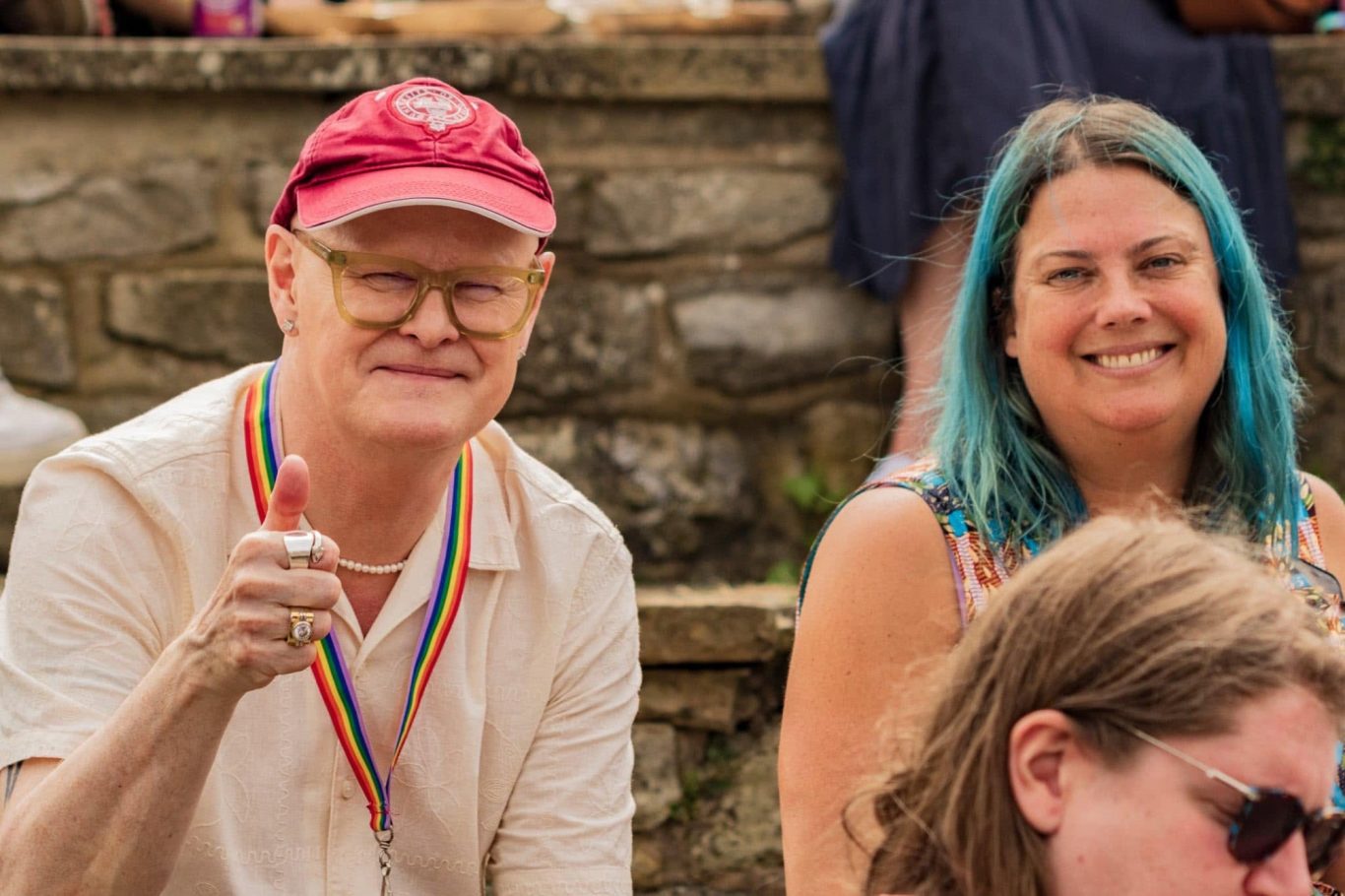 Two smiling individuals, one wearing a red cap and the other with blue hair, at an outdoor event.