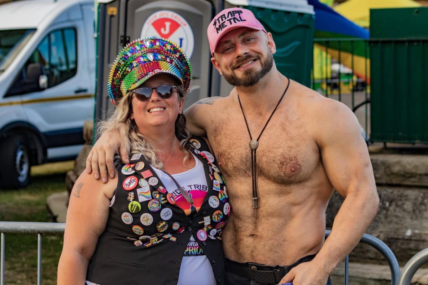 A woman in a colourful hat and a man with a muscular build pose together, smiling.