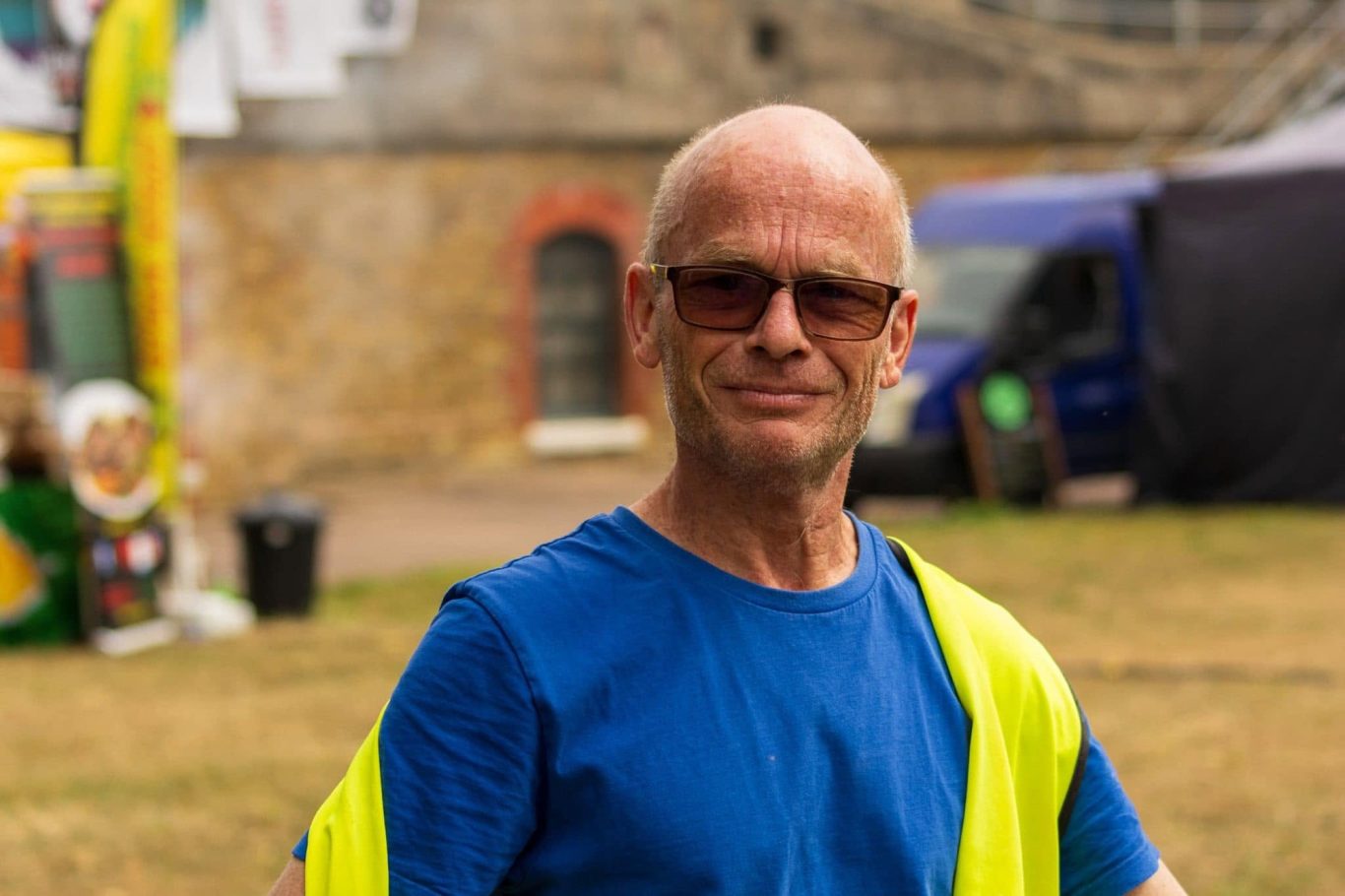 Man wearing sunglasses and a blue shirt, smiling, with a high-visibility vest.