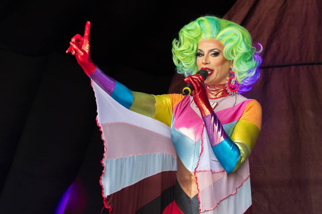 Divina DiCampo - 2024 A performer in vibrant drag attire with colourful hair, gesturing while singing on stage.