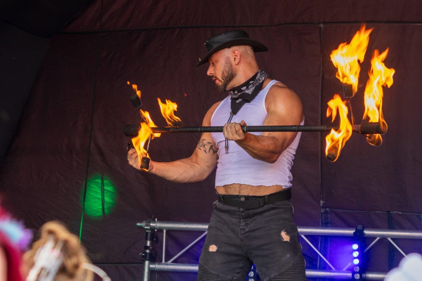 Marshall Arkley - 2024 A performer in a hat juggling fire while wearing a sleeveless top and trousers.