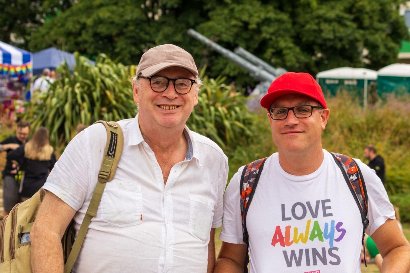 Two smiling men pose together outdoors, one in a white shirt and the other in a red cap.