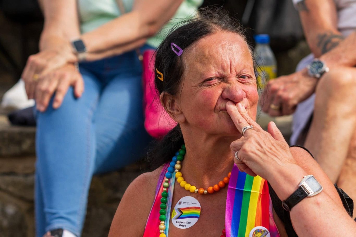 A woman with a rainbow necklace gestures playfully while sitting among a crowd.