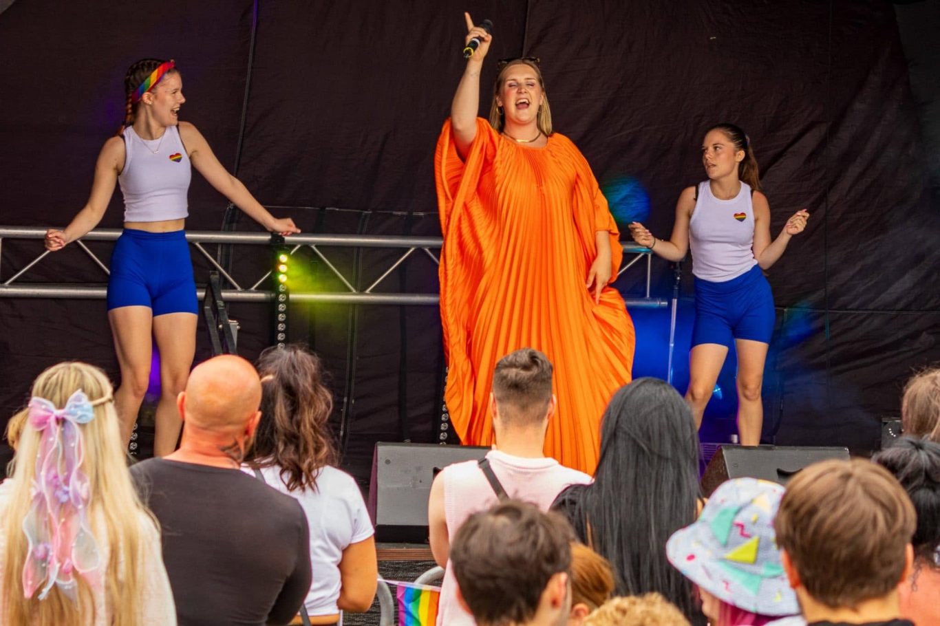 GYG - 2024 A performer in an orange outfit sings on stage, flanked by two dancers in blue shorts.