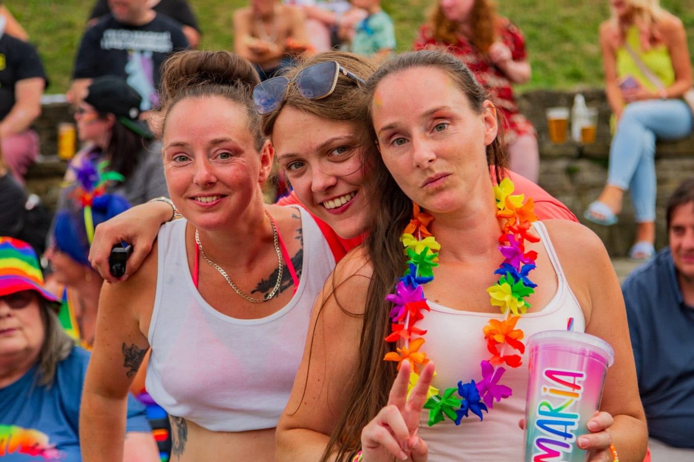 Three women posing together, wearing colourful leis, with a festive crowd in the background.