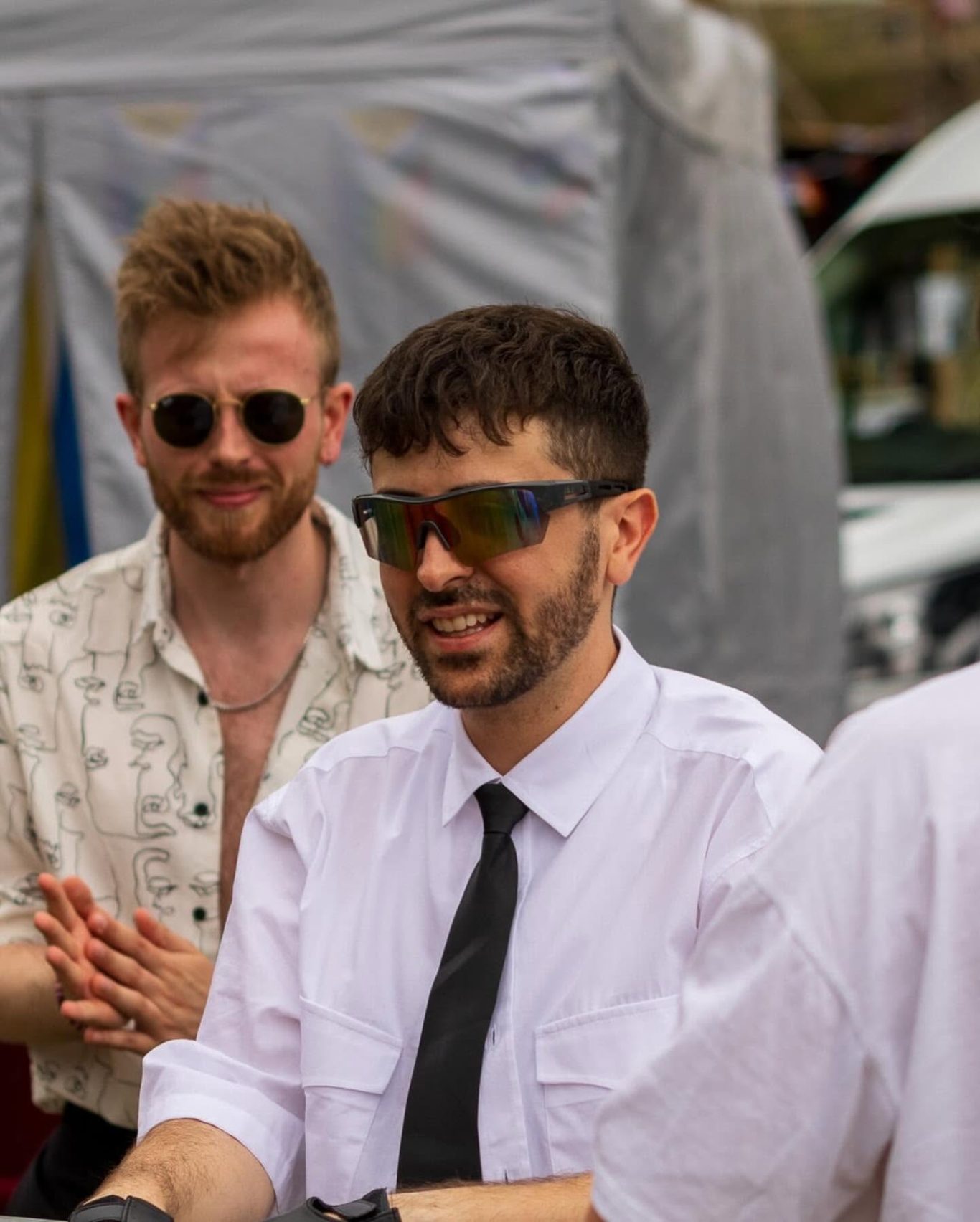 Two men, one in sunglasses and a white shirt, the other in a suit and tie, interacting.