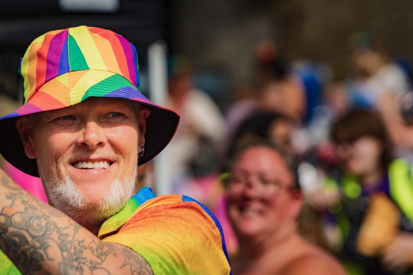 Man in a rainbow-patterned bucket hat and matching shirt, smiling at a lively event.