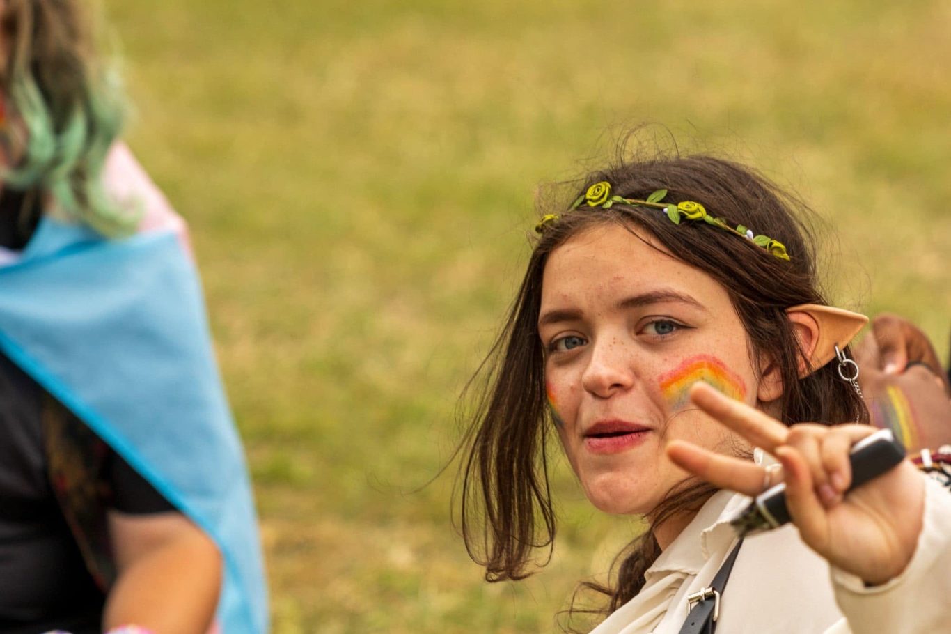A young person with pointed ears and a flower crown flashes a peace sign, smiling.