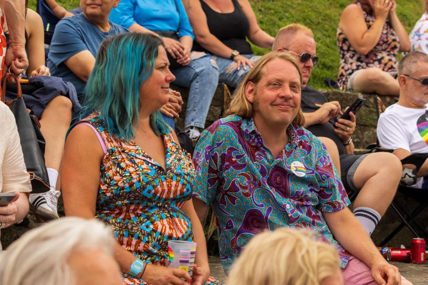 A woman with blue hair and a patterned dress sits next to a man in a vibrant shirt.
