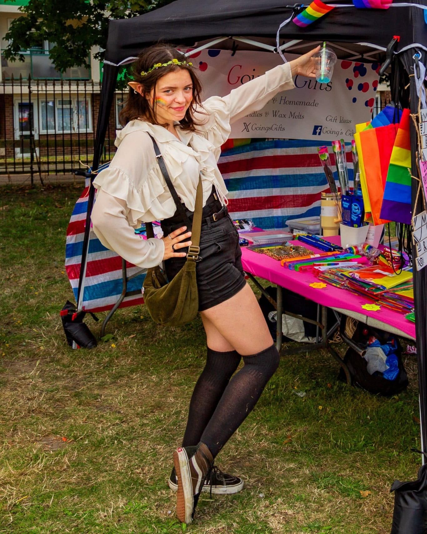 Person in a casual outfit posing by a colourful stall decorated with rainbow flags.