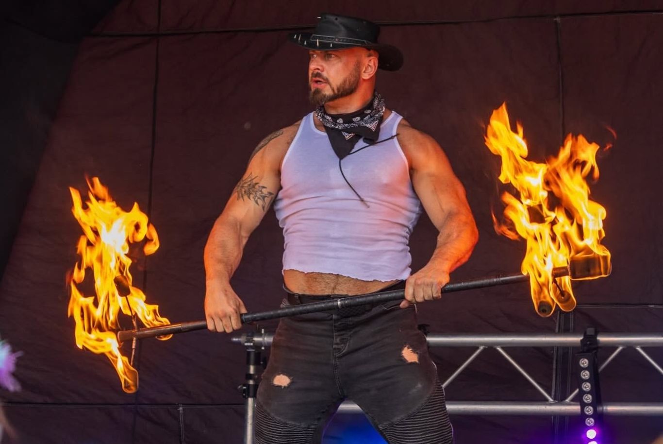 Marshall Arkley - 2024 A muscular performer skilfully twirls flaming torches on stage.