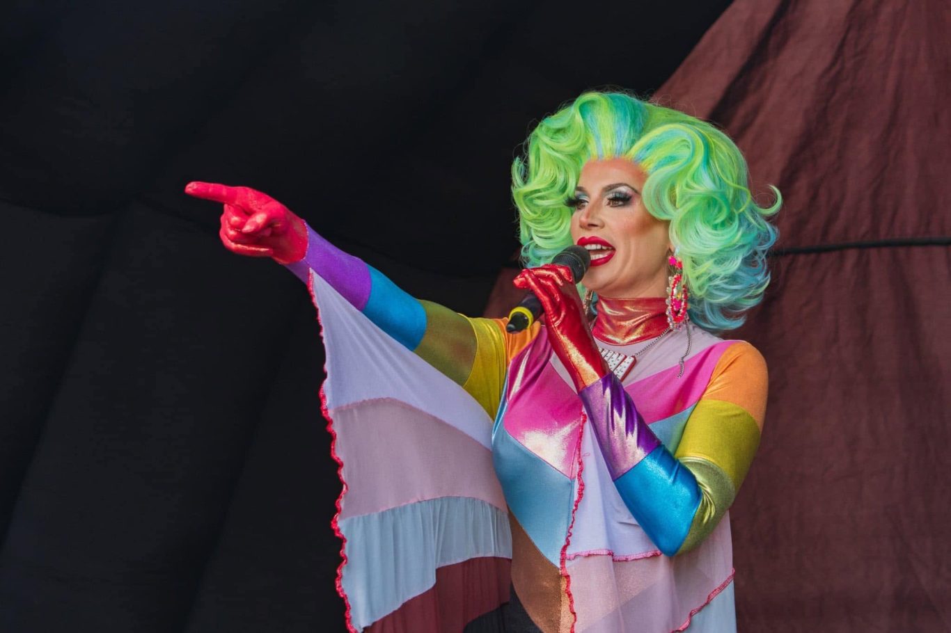 Divina DiCampo - 2024 Performer in vibrant rainbow costume with green curly hair, singing into a microphone.
