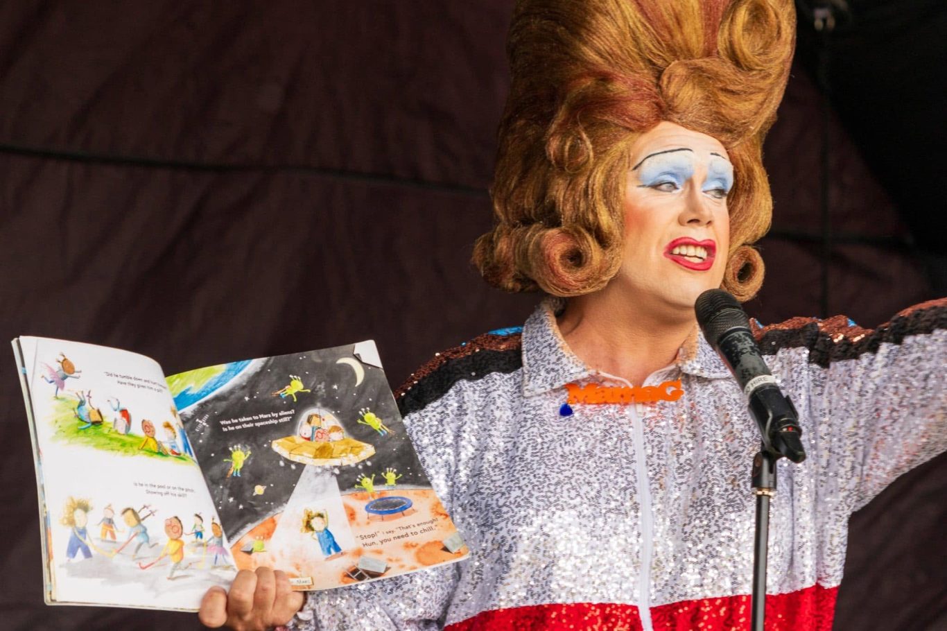 Mama G - 2024 A performer in a sparkly outfit reads from a colourful children's book on stage.