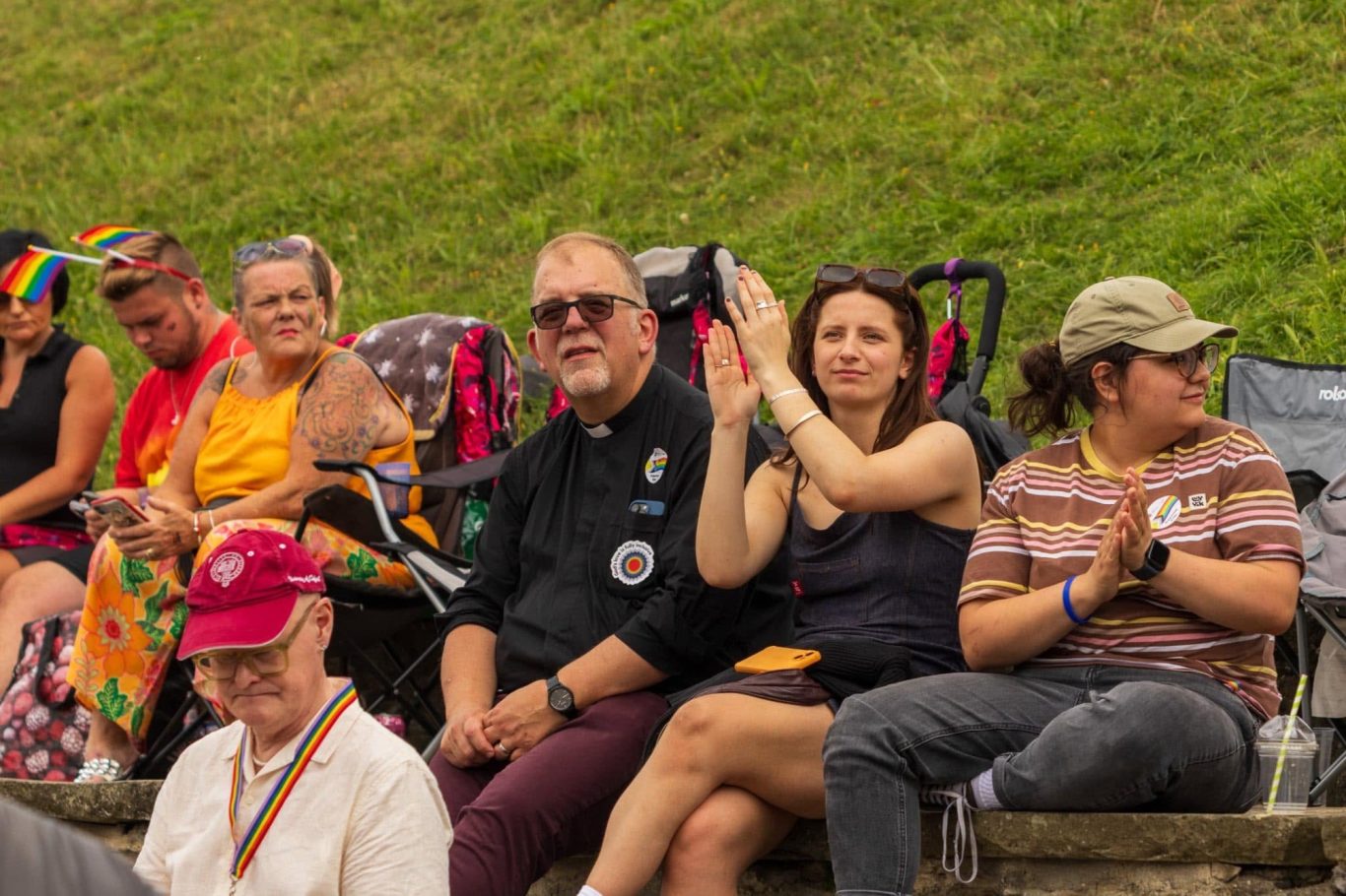 People sitting on a grassy area, some clapping and others taking photos, during a celebration.