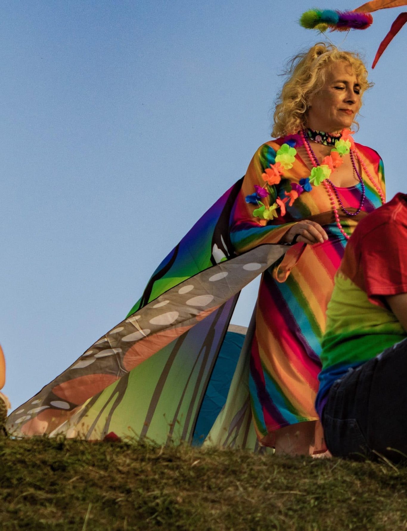 A person in a vibrant rainbow outfit standing on grass, surrounded by colourful decorations.