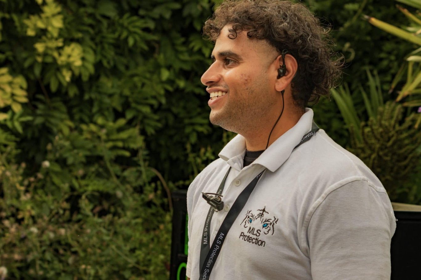Man with curly hair, wearing a white shirt, smiling against a lush green background.
