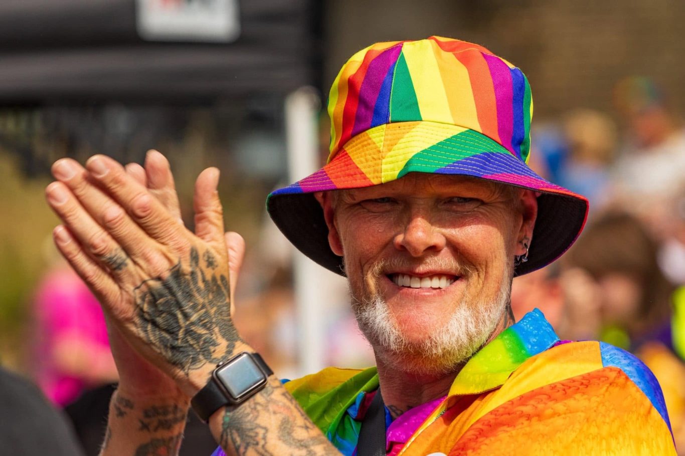Smiling person in a rainbow hat and shirt, clapping in a festive atmosphere.