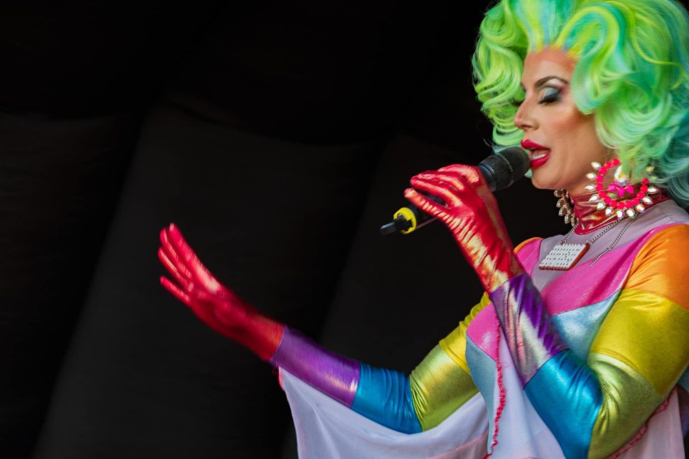 Divina DiCampo A drag performer with bright green hair and colourful attire gestures while singing.