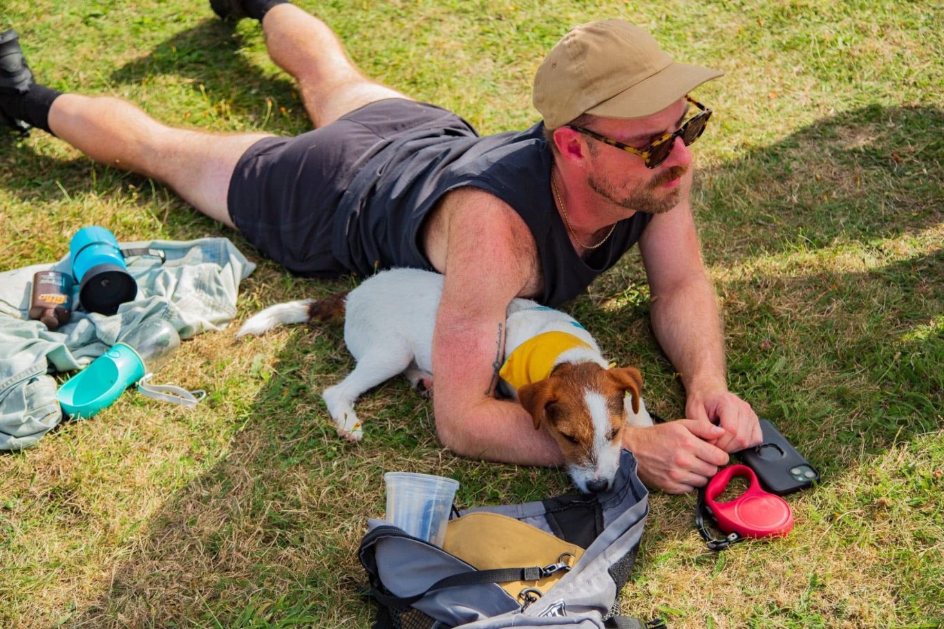 Man in a cap and sunglasses lying on grass with a small dog beside him, both relaxed.
