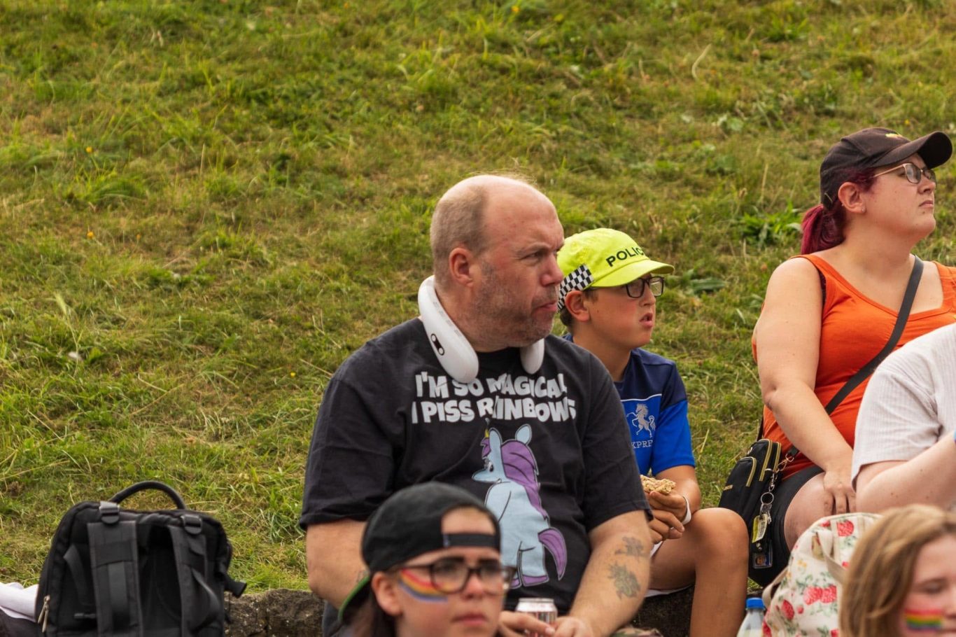 A man in a black T-shirt sits on grass, with a boy and other people in the background.