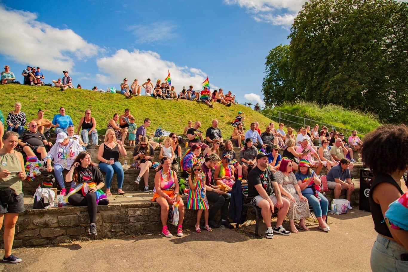 Crowd of people sitting on grassy steps, enjoying a sunny outdoor event with flags.