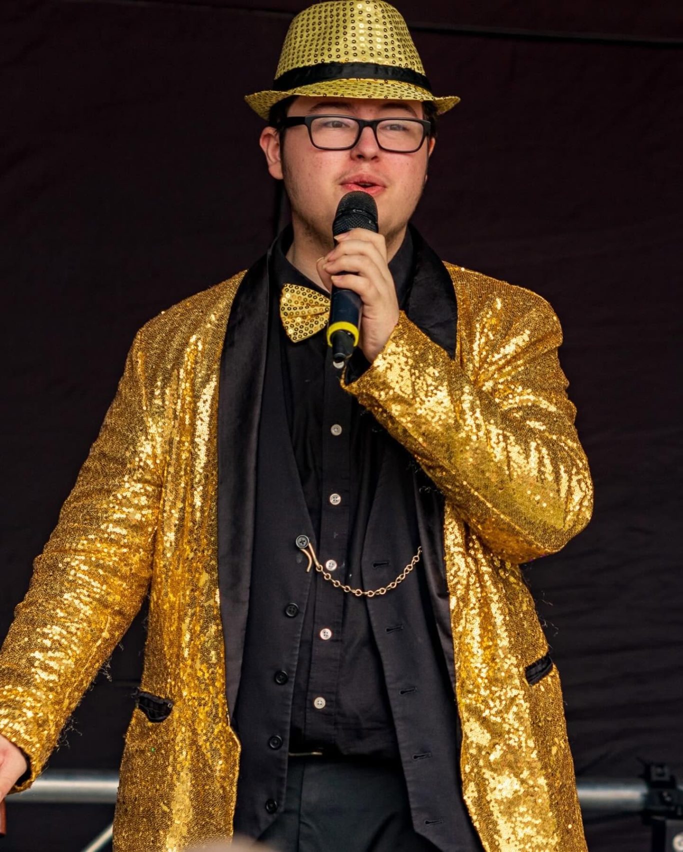GYG - 2024 Person wearing a gold sequined jacket and hat, holding a microphone while performing.