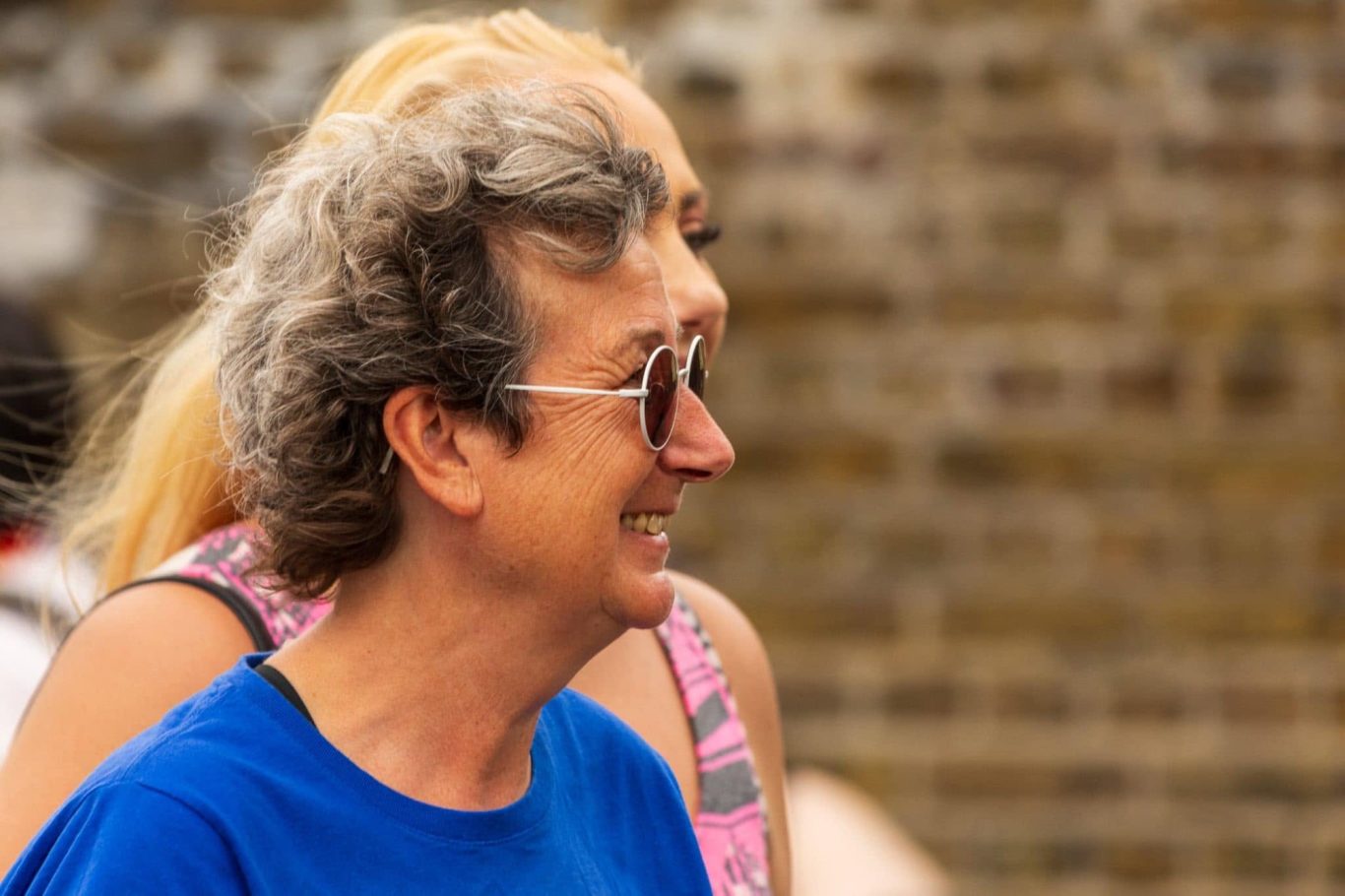 Smiling person with curly hair and sunglasses, in profile, with another person in the background.