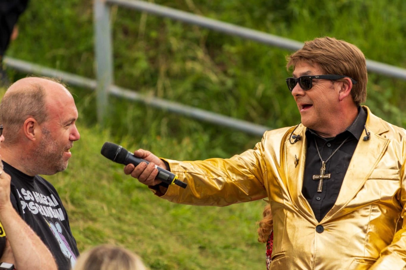 Elton John Tribute A man in a gold jacket and sunglasses speaks into a microphone to another man.