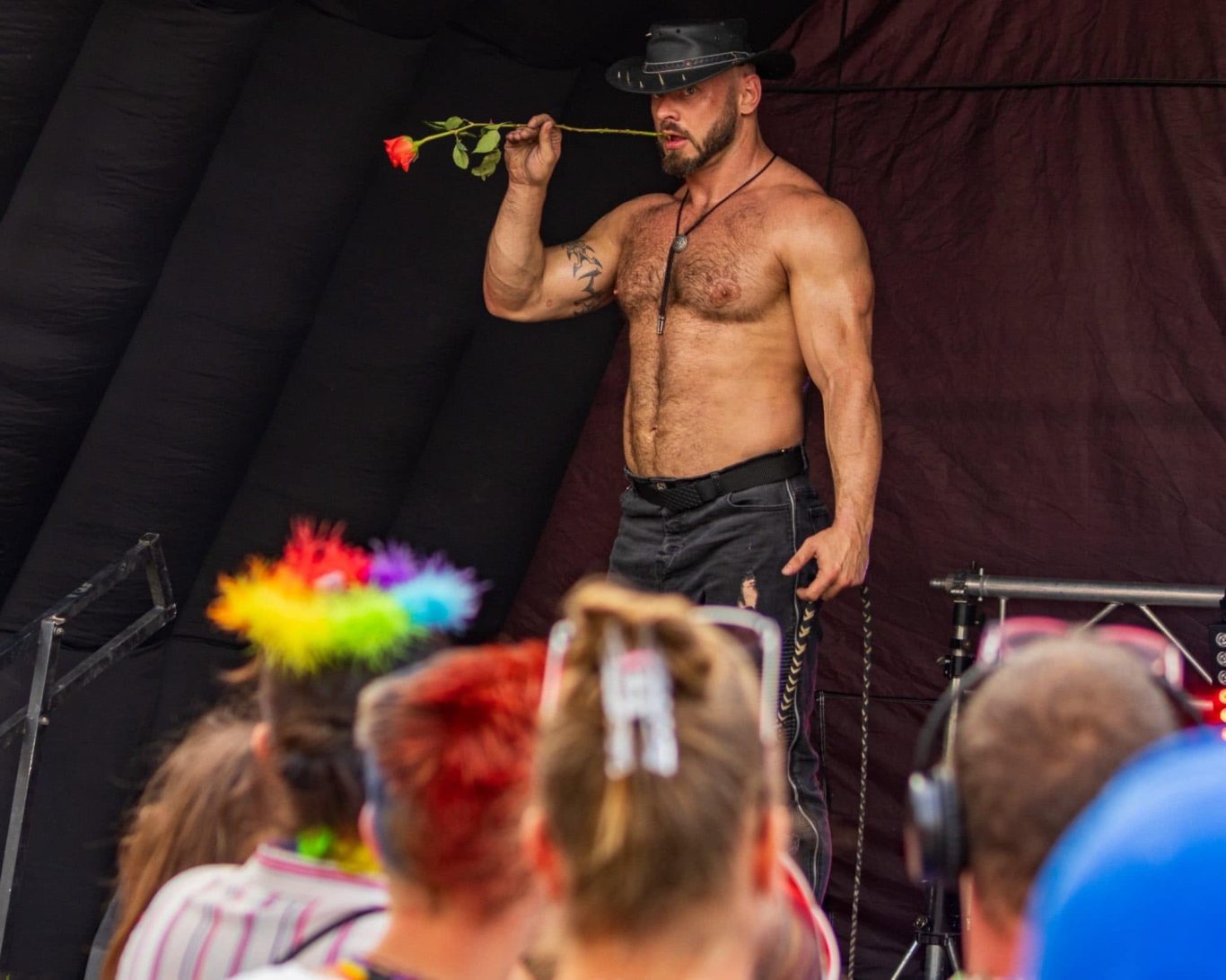 Marshall Arkley - 2024 Muscular performer in a black hat holding a rose, with a colourful crowd in the foreground.