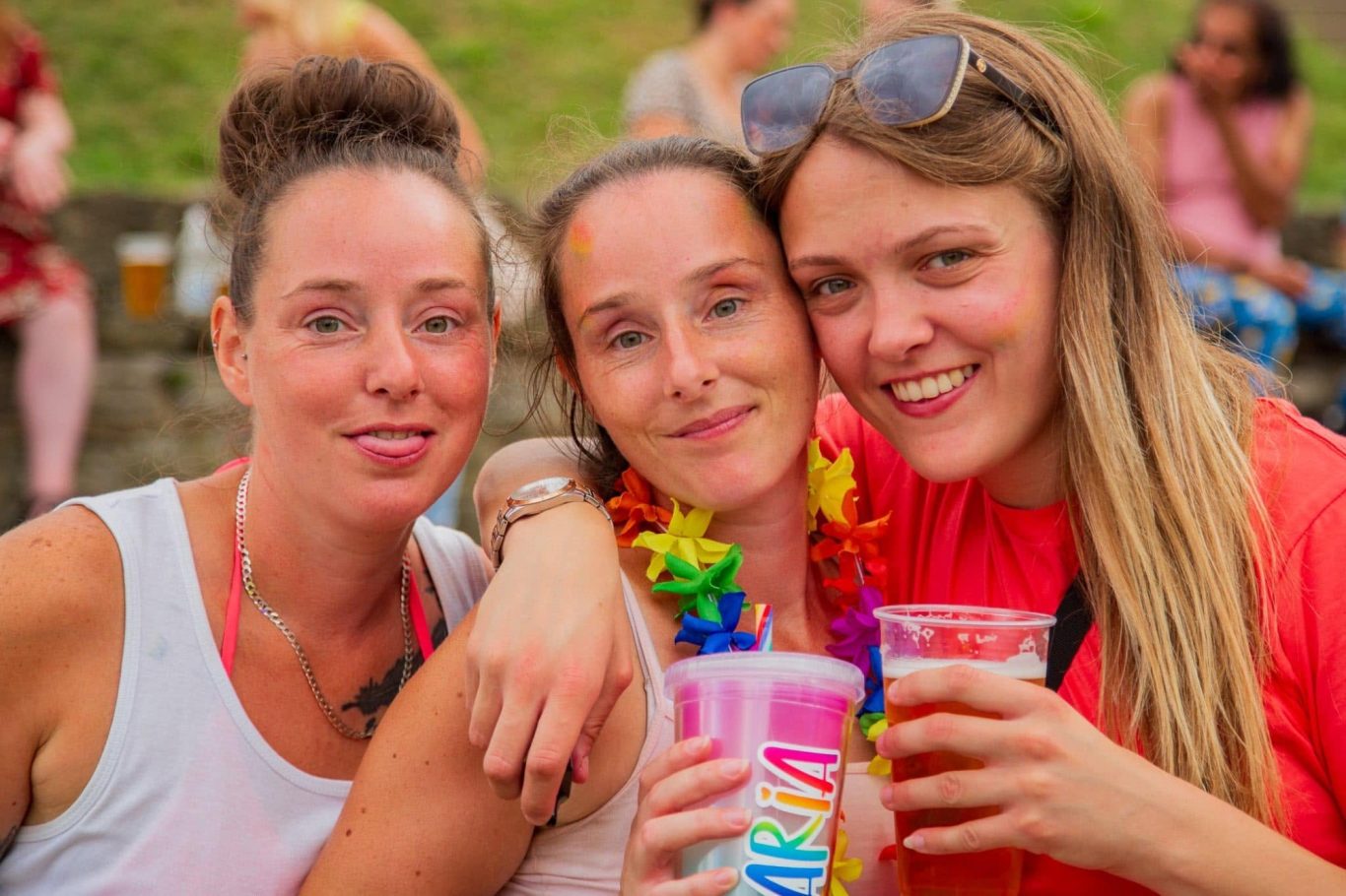 Three women smile and pose together, each holding colourful drinks, in a festive outdoor setting.