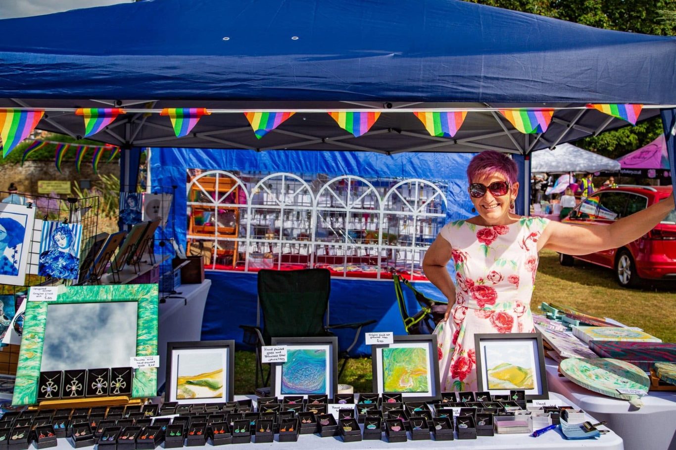A woman in a patterned dress stands behind a stall featuring colourful art prints and crafts.