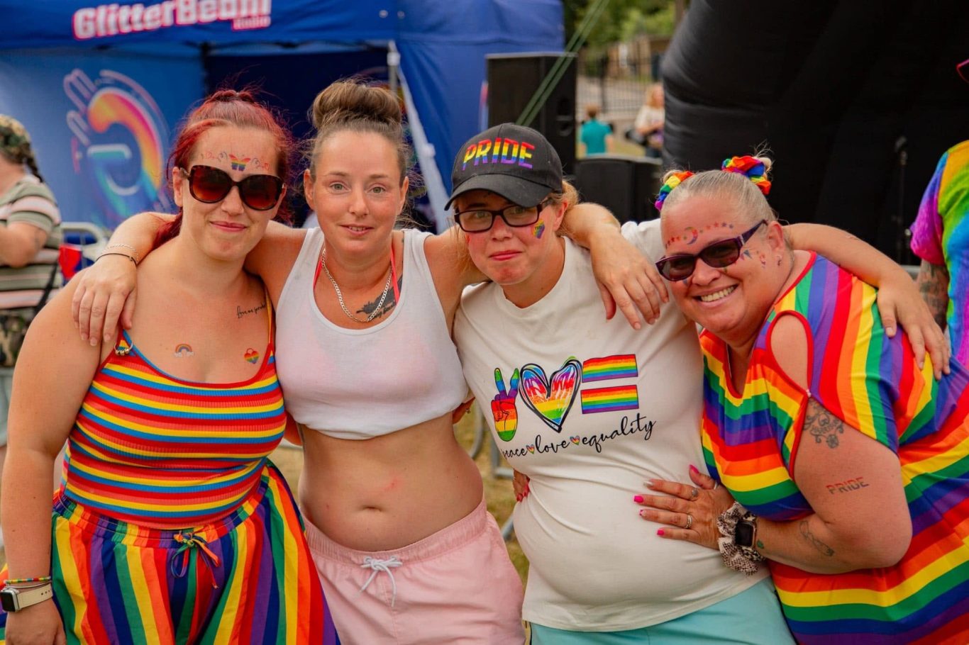 Four people wearing bright rainbow clothing, smiling and celebrating at a Pride event.