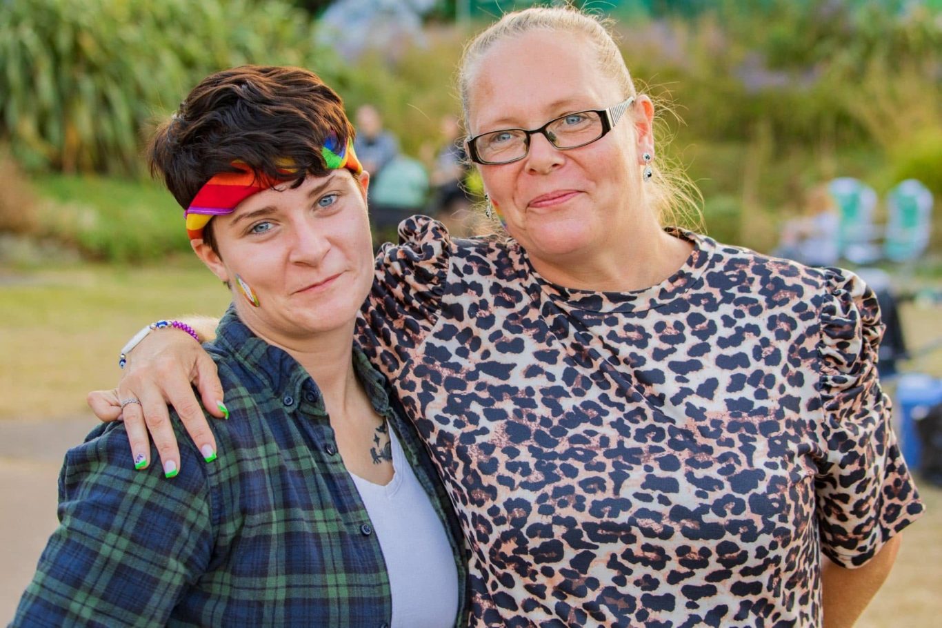 Two women pose together outdoors, smiling, with a vibrant background of greenery.