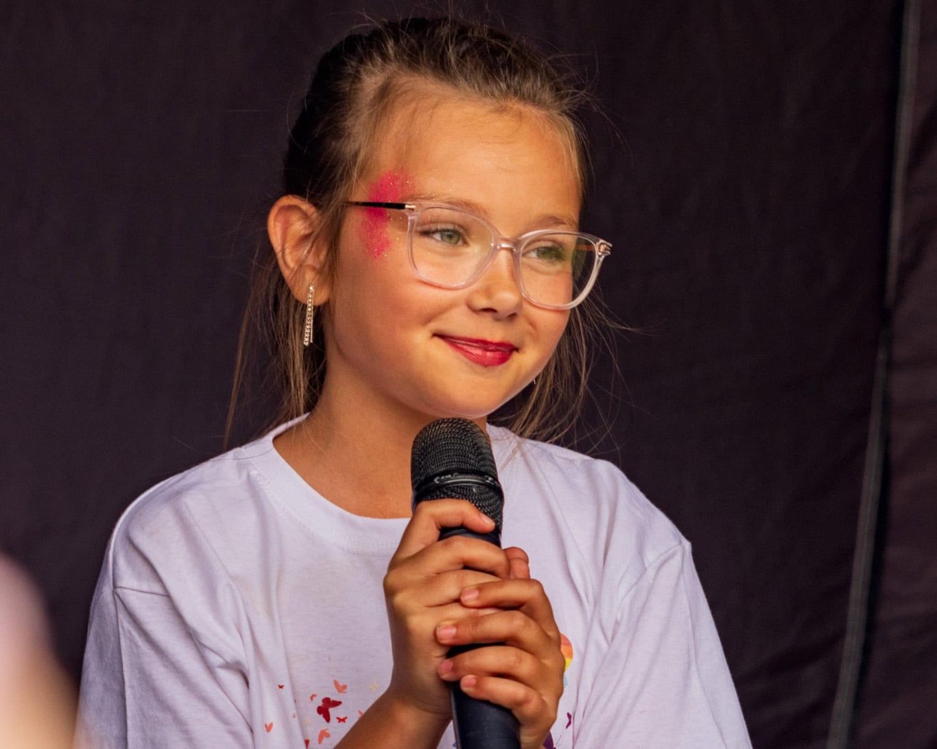 GYG - 2024 Young girl with glasses holding a microphone, smiling on stage.