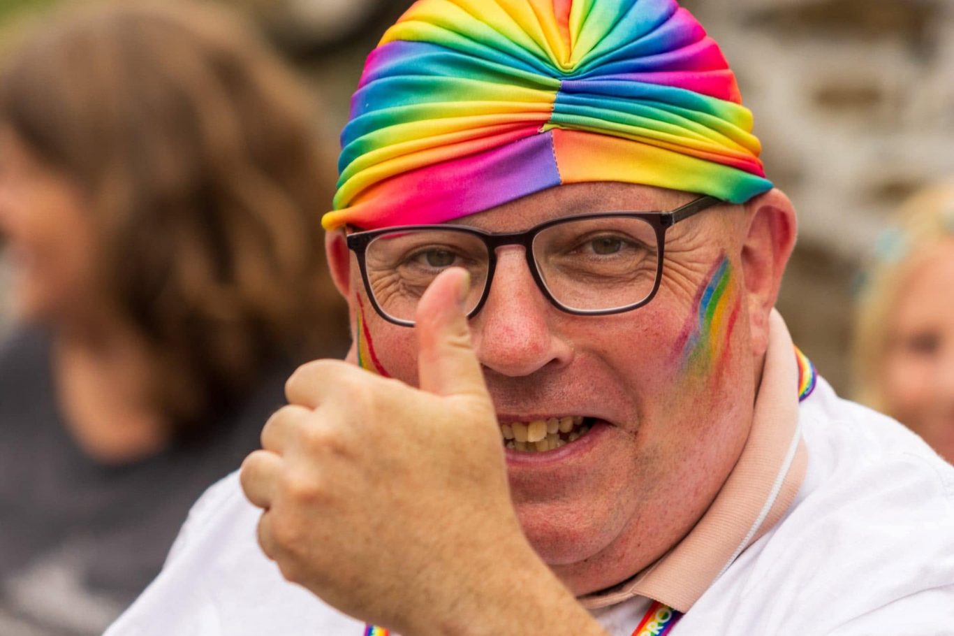 Man with glasses and rainbow headband, giving a thumbs-up, celebrating pride.