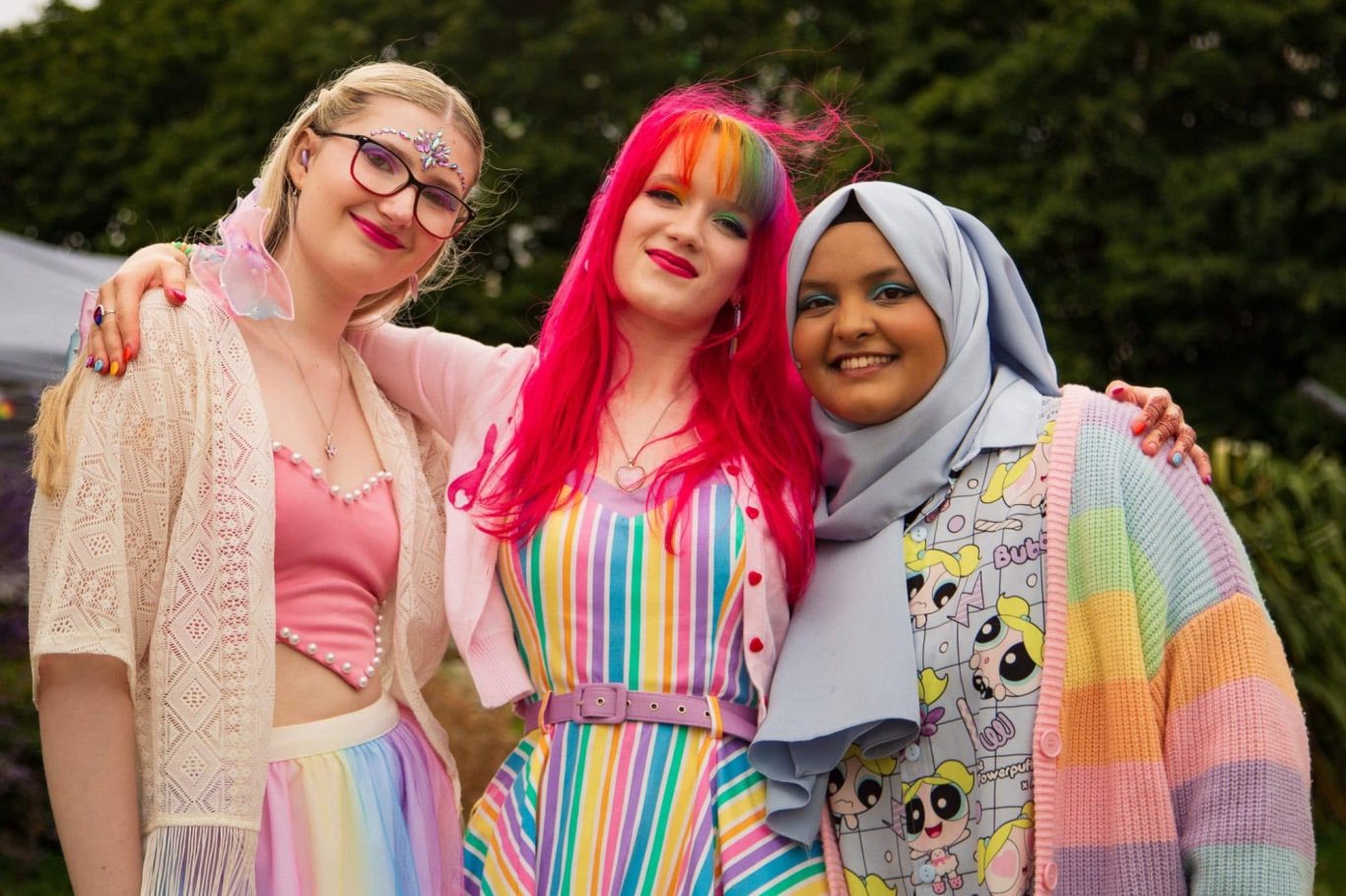 Three women pose together, wearing colourful outfits and smiling in an outdoor setting.