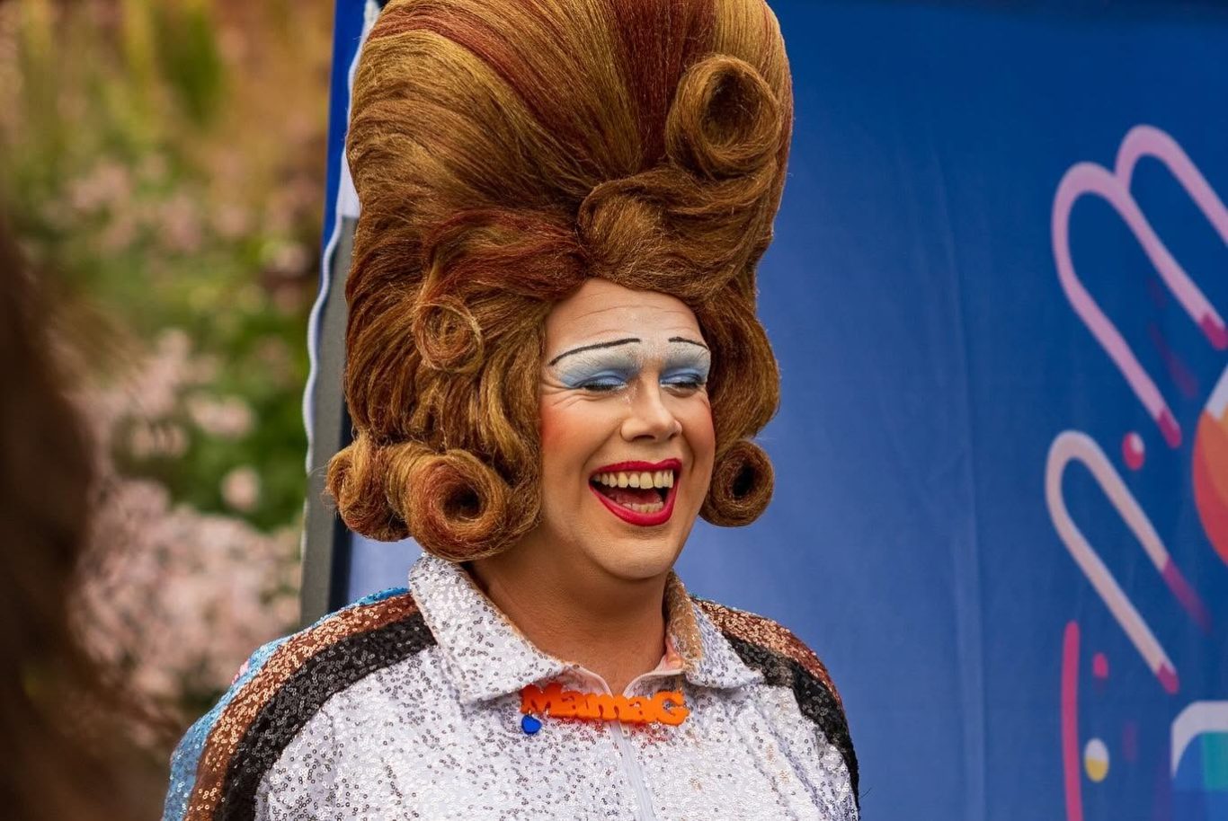 Mama G - 2024 Person with a large styled wig and bright makeup, smiling against a colourful background.