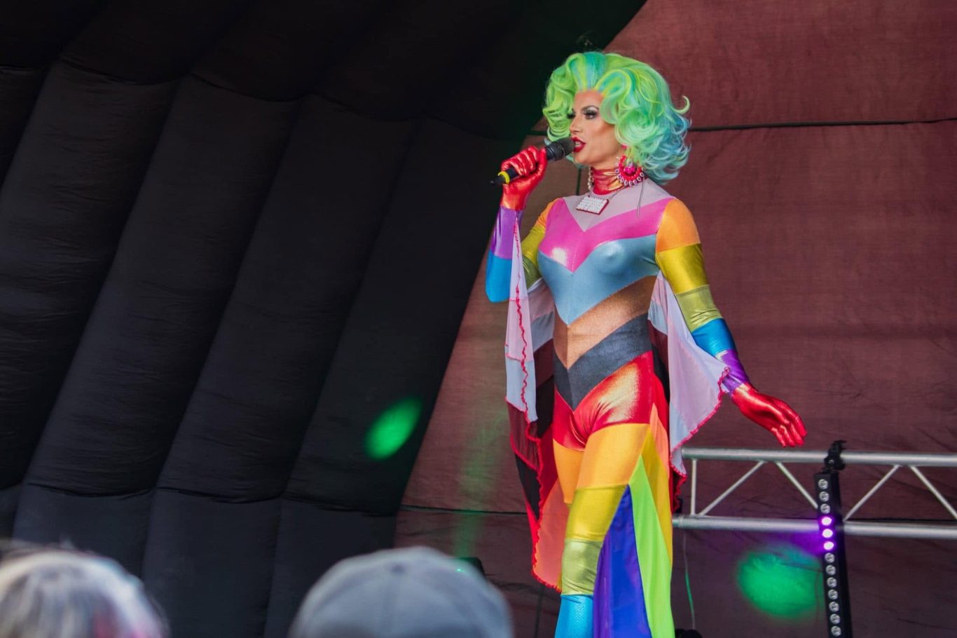 Divina DiCampo - 2024 A performer in a colourful, rainbow-themed costume sings on stage.