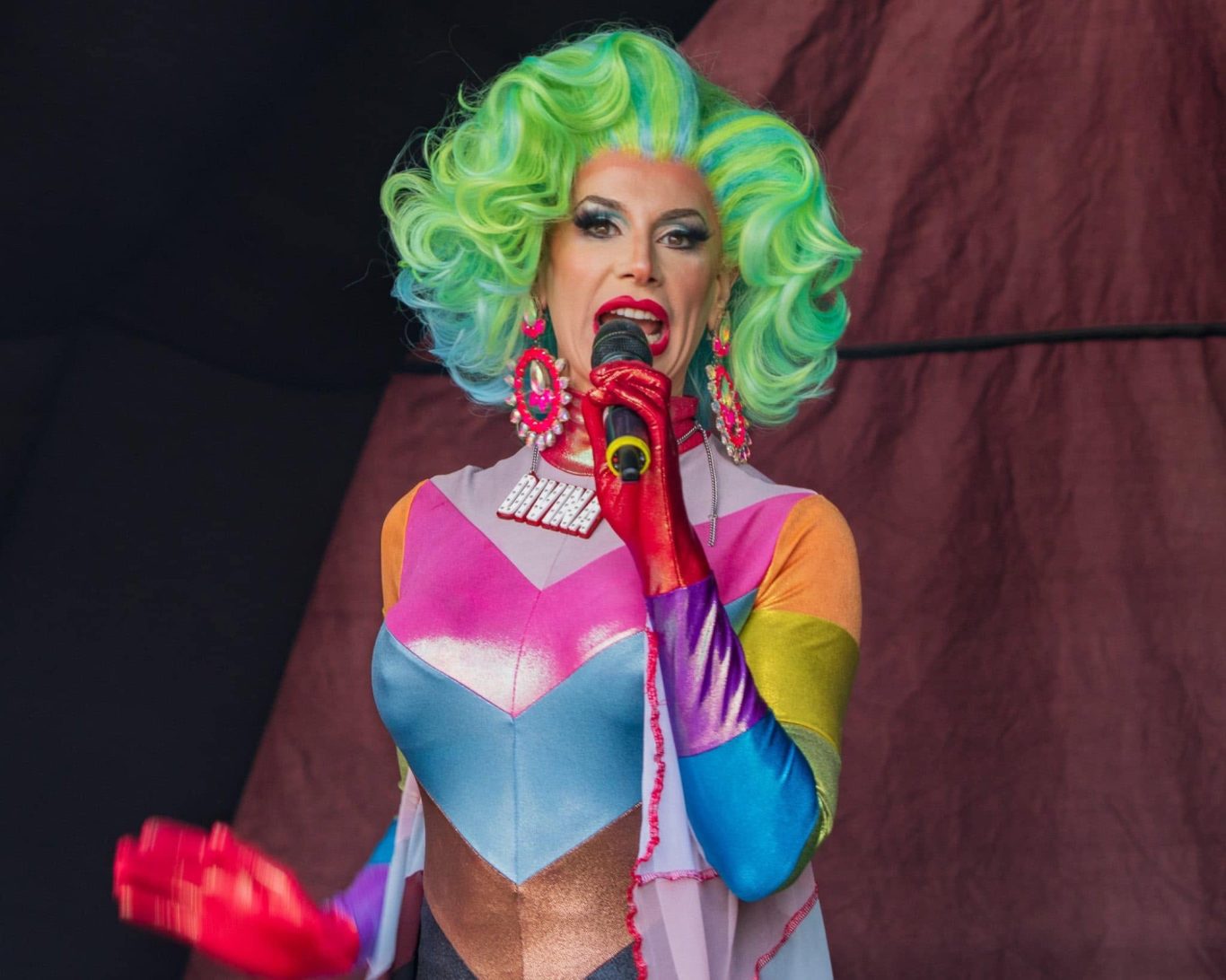Divina DiCampo - 2024 A performer in vibrant, multi-coloured attire with green hair and dramatic makeup speaks into a microphone.