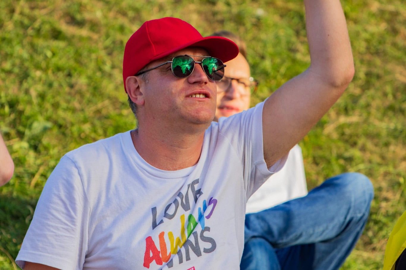 Man in a red hat and sunglasses, wearing a white T-shirt, raising his hand outdoors.