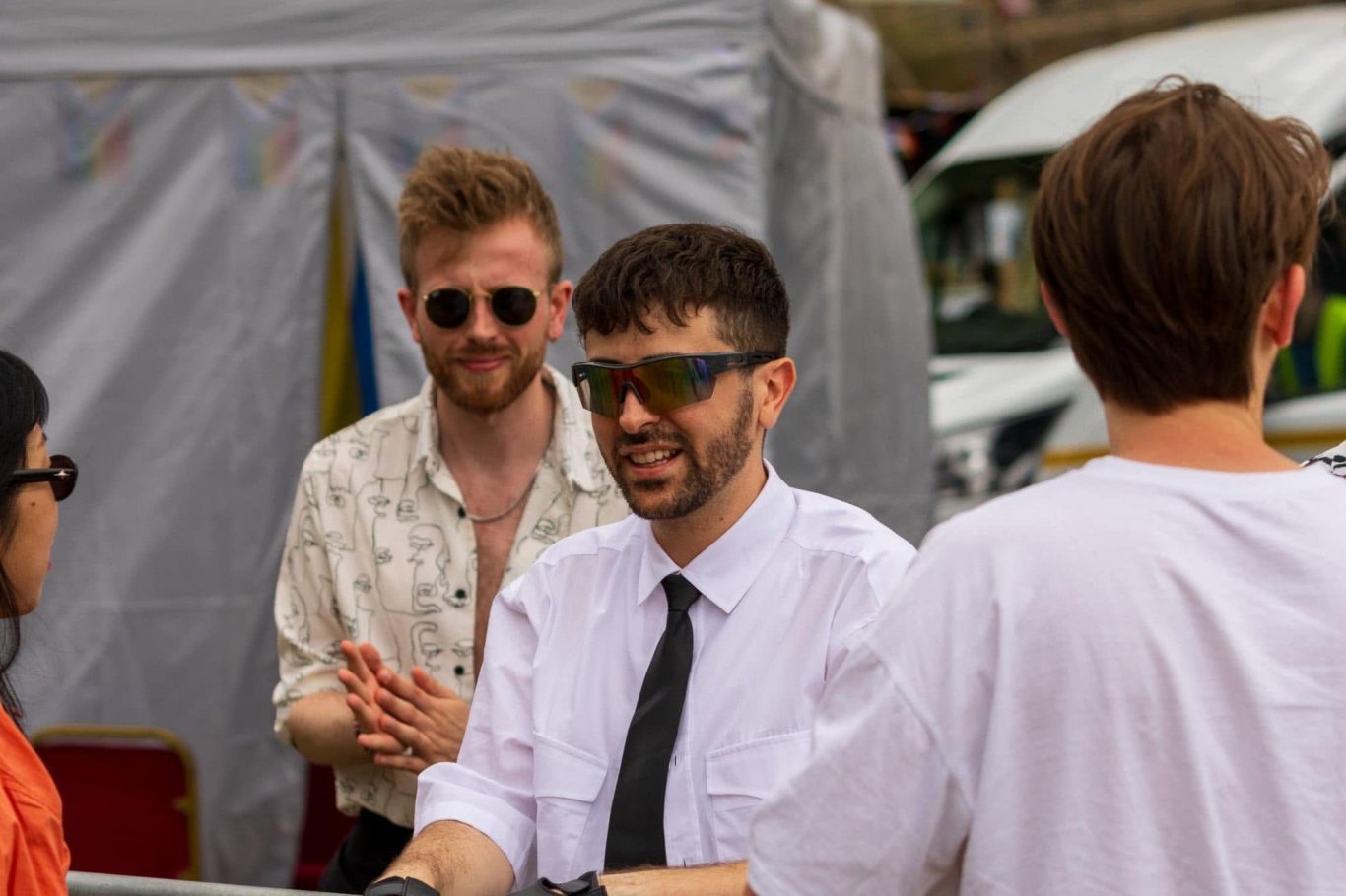 Two men in sunglasses are interacting with a group, outdoors at a casual event.
