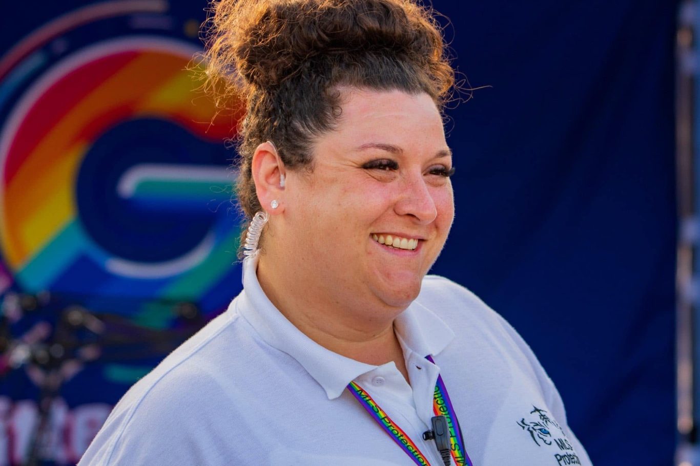 A smiling woman in a white shirt with a rainbow badge, set against a colourful backdrop.